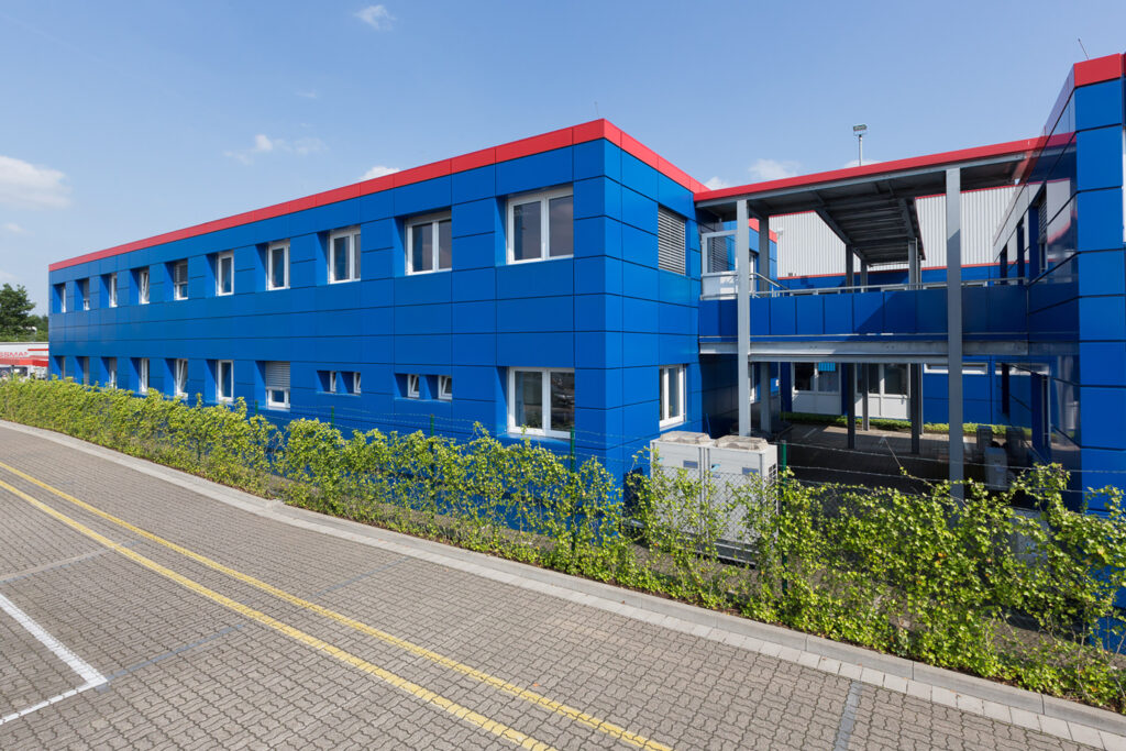 Verwaltungsgebäude Schmitz Cargobull AG 1 Außenansicht eines blauen Bürogebäudes in Modulbauweise mit einem begrünten Zaun davor